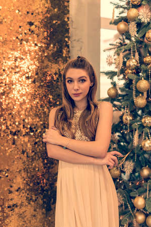 girl in a shiny evening dress. The girl is dreaming. Christmas Eve. Cozy holiday at the fur-tree with lights and gold decor. girl in a shiny dress on a background of Christmas background in the studioの写真素材