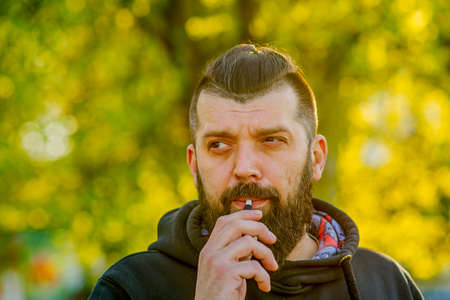 serious hooded guy with beard sunny outdoor on blurred background. Man smokes electronic smokes