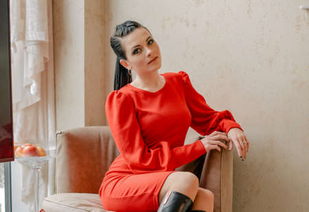 Young girl in a red dress indoors. model. Emotions of a modern lady. The girl is sitting in a chair