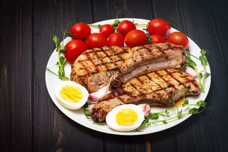 Two grilled pork steaks on a white plate with sprouted chickpeas, cherry tomatoes and boiled egg.の写真素材