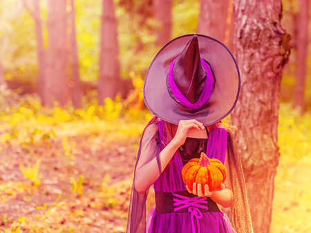 A girl in a Halloween costume with a hat over her eyes with orange pumpkin in the forestの写真素材