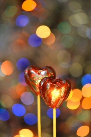 Chocolate candies for valentine's day. Red heart shape chocolate on lolly stick. Chocolate heart in red foil on a stick on a bokeh blurred backgroundの写真素材