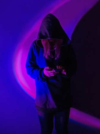 Unrecognizable faceless person in dark hoodie holding phone on bright multicolored neon glowing...