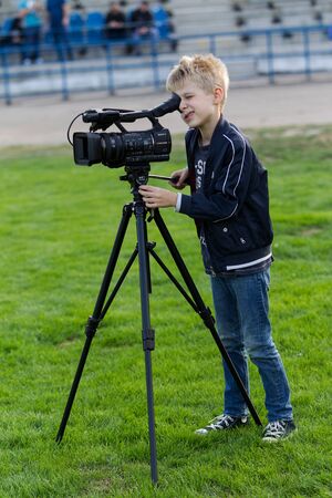 ODESSA, UKRAINE - CIRCA 2019: A little boy, a teenager, shoots 4K video on a professional video camera on the green field of the stadium. Young videographer with a television cameraのeditorial素材