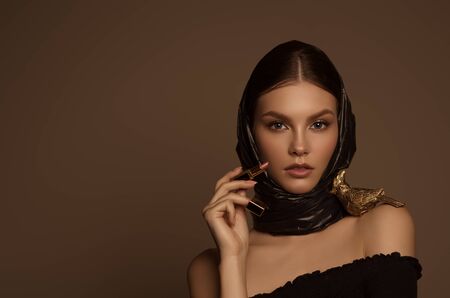 Beautiful girl in a scarf holds lipstick in her hands on a brown background.の写真素材