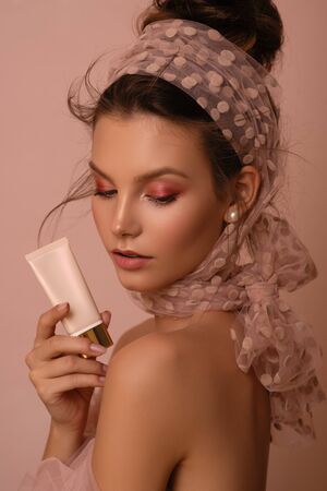 Beautiful young woman with perfect skin in a scarf on a pink background holds cosmetics in her hands.の写真素材