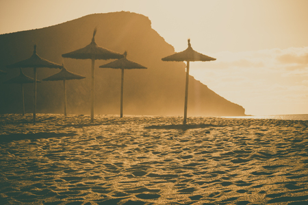 Thatch palapa umbrellas against sunlight at beach Playa la Tejita, Montana roja on background. Tenerife island, Spain. Retro toned imageの写真素材