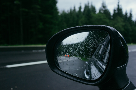 Rain drops on car side view mirror and car lights reflection. Road and forest on backgroundの写真素材