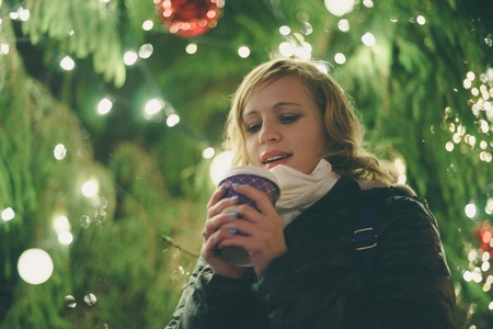 Happy young lady with cup of coffee against illuminated christmas tree, shallow depth of fieldの写真素材