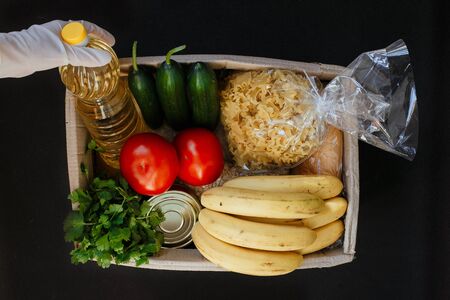 A hand in a rubber glove holds tomato. A donation box with a food. Fruits, vegetables, canned food, pasta and sunflower oil in a box. Social assistance with food.の写真素材