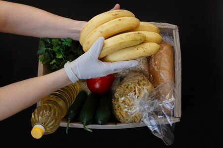 Valanter's hand in protective gloves fill the box with food on a black background.Donation concept. Voluntary assistance to those in need.の写真素材