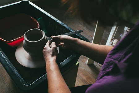 Woman potter in apron sculpts a deep bowl from the clay on a potter's wheel and cuts off excess clay...