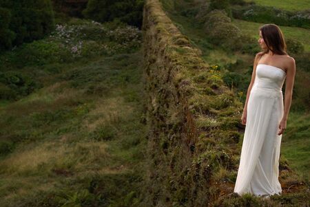 Portrait of young bride in white dress standing alone on grassy brickwall. Background of romantic green field and hillsの写真素材