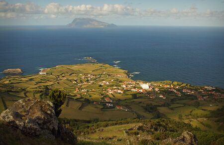 Beautiful sunny view of Sao Miguel, Azores island. Green fields on hills, cosy village, small houses with red roofs. Background of bright blue ocean water and neighbour island with mountainsの写真素材