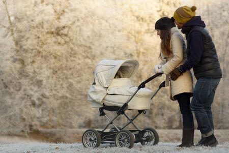 Young parents in winter clothes looking at child in white baby stroller. Backgroung on sunny snowy winter park and frosty icy trees, childcare and loveの写真素材