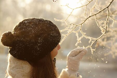 Sunny portrait of young woman in black hat with pompom and white mittens touching snowy icy tree branch in winter forestの写真素材