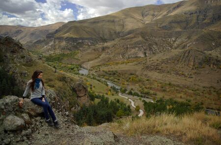 Young slim woman with long hair sitting elegant on stone on background of mountains and brown valley with river, view from sideの写真素材