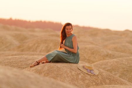 Young beautiful woman with long hair in blue dress sitting elegant and thoughtful on sand at sunrise in sandy desertの写真素材