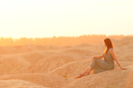 Young beautiful woman with long hair in blue dress sitting on sand at sunrise in sandy desert. Looking thoughtful downの写真素材