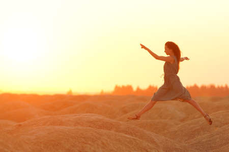 Elegant woman in long dress with straw hat junping elegant on sandy hills in desert. Sunrise,...
