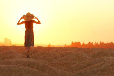 Elegant woman in long dress with straw hat standing on sand in desert, back view. Sunrise, backlight photoの写真素材