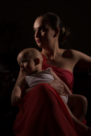 Beautiful woman with her child in red evening dress in dark environmentの写真素材
