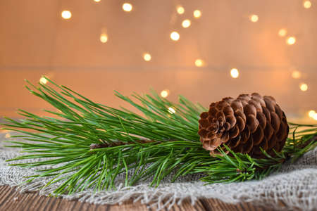 A Christmas cone and a branch of a fir tree on a wooden table against the background of lights of...
