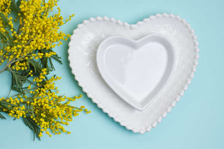 Two ceramic plate in the shape of a heart and mimoza flowers on a blue background, top view. Love. Tableware. Postcard, frame, banner.の写真素材