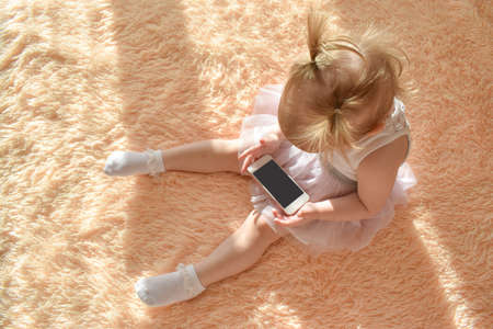 A little girl sits on the bed and looks at the phone. Dependence on gadgets.の写真素材