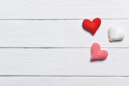The background of Valentine's Day. Small knitted hearts on a white wooden background. Hearts of three colors. Minimalism is love. Copy space. Flat lay, top view.の写真素材