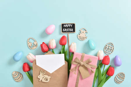 A bouquet of tulips with a postcard, a gift and wooden Easter eggs with a plaque on a light blue...