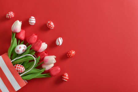A beautiful bouquet of tulips in a paper eco-bag, with painted Easter eggs on a red background. Banner for a happy Easter holiday. Space for copying. Flat position, top view.の写真素材