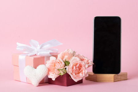 Smartphone on the background of a pink gift box with roses and a knitted white heart on a light pink background with a place for text.の写真素材
