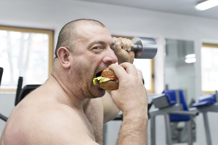 A big fat hungry man eats a hamburger with meat and cheese in the gymの写真素材