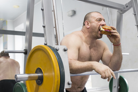 A big fat hungry man eats a hamburger with meat and cheese in the gymの写真素材