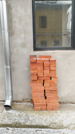 the wall of the house has a window and a drainpipe, bricks are stacked next to it. The window...
