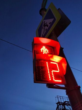 a red traffic light for pedestrians on a pole with a pedestrian crossing sign at dusk against a blue sky from bottom to topの写真素材