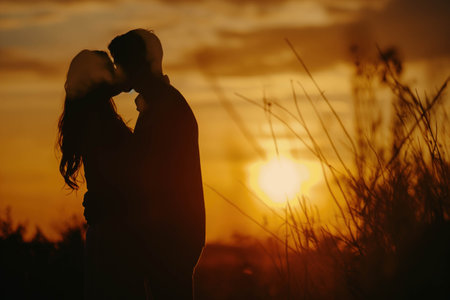 Silhouette of a young couple in love kissing on background of the orange sky during sunset.の素材