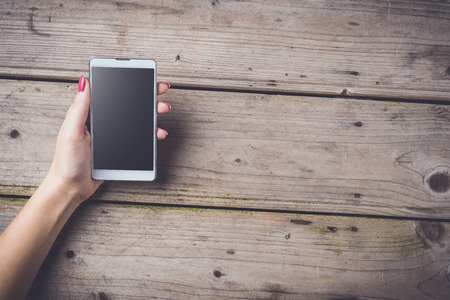 Woman holding white smart phone over wooden tableの写真素材