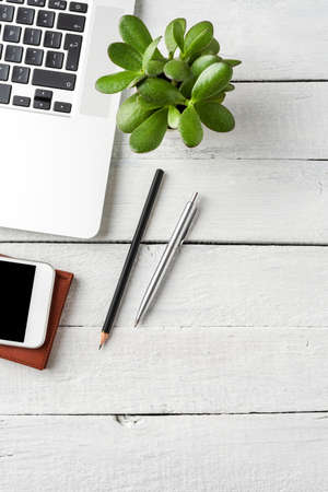 Laptop, mobile phone, notebook, pen and small flower on white wooden desktop. Business background...