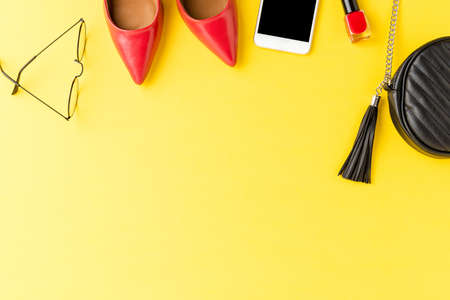 Fashion background. Red high heels, purse, phone, eyeglasses and nail polish on yellow table. Flat...