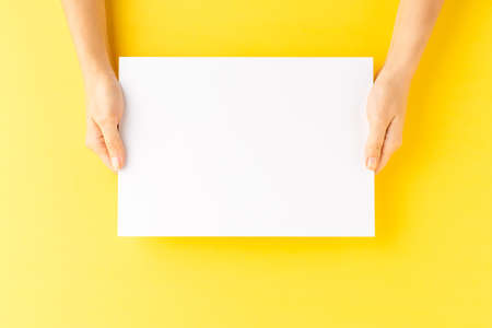 Overhead shot of woman's hands holding blank paper sheet on yellow table. Close up