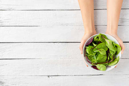 Overhead shot of female hands holding green salad in bowl on white wooden background with copyspace