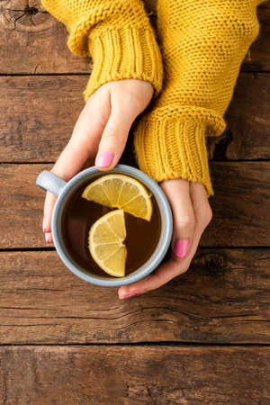 Overhead shot of female hands knitted sweater holding cup of tea with lemon on wooden tableの写真素材
