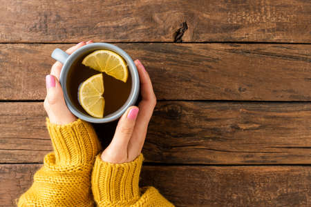 Womanâs hands in knitted sweater holding mug of tea with lemon on wooden background with copyspace. Top viewの写真素材