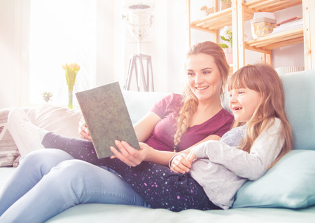 Mother and daughter sitting on sofa at home and reading book togetherの写真素材