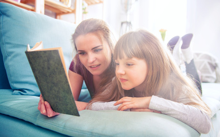 Mother and daughter sitting on sofa at home and reading book togetherの写真素材