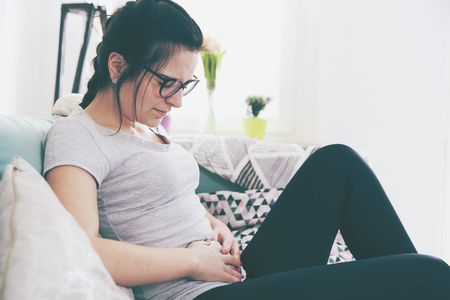 Young woman in pain while sitting on comfortable sofa, home interiorの写真素材