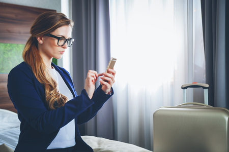 Pretty business woman with suitcase in modern hotel room using laptop and smartphoneの写真素材