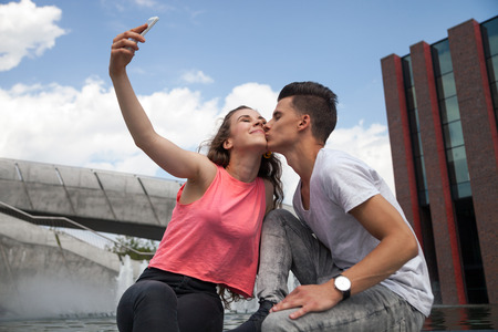 Young couple giving kiss sitting in town outdoor with mobile phone taking selfieの写真素材
